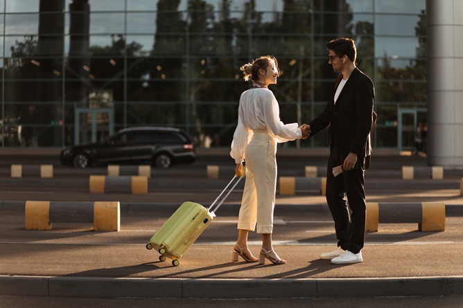 happy blonde woman white blouse beige pants holds yellow suitcase greets with man black suit near airport
